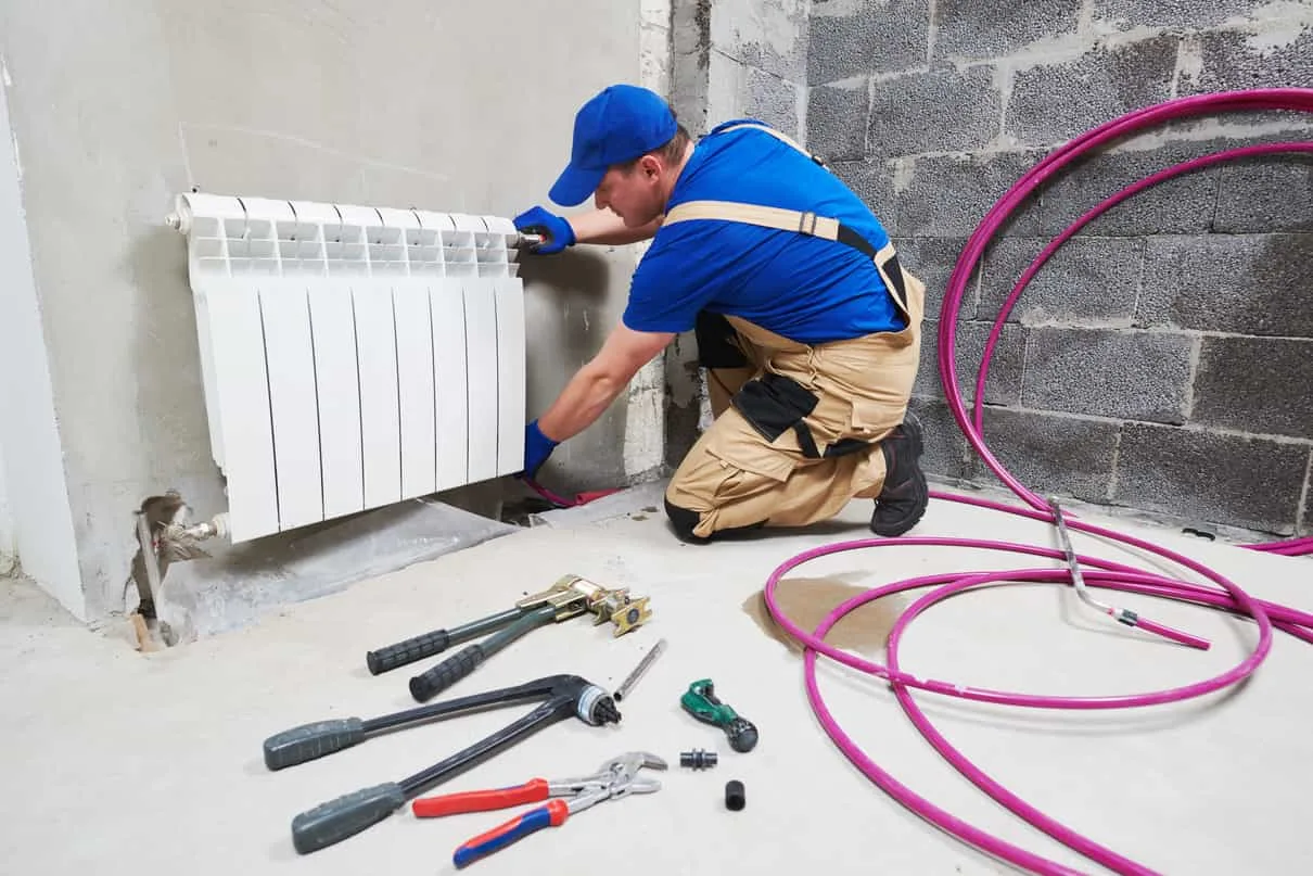 Instalator w niebieskiej czapce montuje biały grzejnik członowy, na podłodze narzędzia i rury