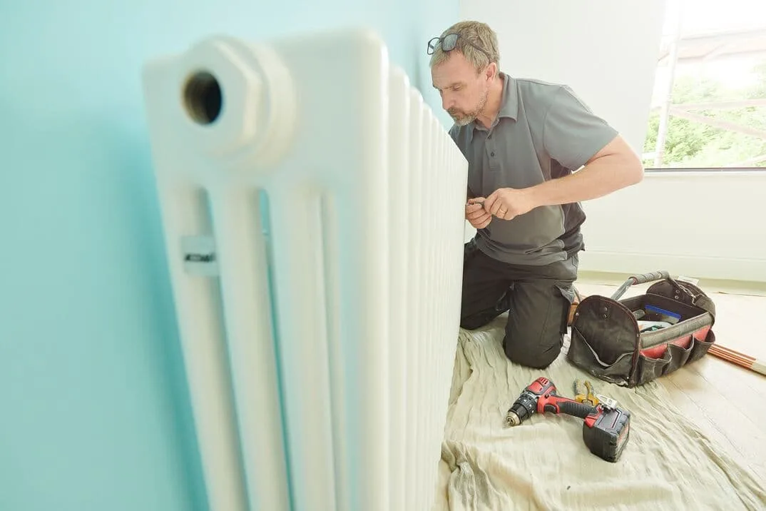 Instalator klęczy przy białym grzejniku kolumnowym, obok wkrętarka i torba z narzędziami
